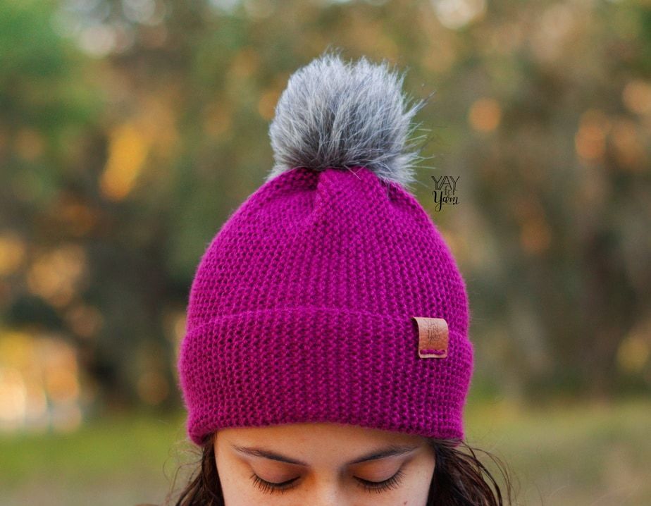 dark purple hat in sport weight yarn with grey faux fur pom pom and leather tag