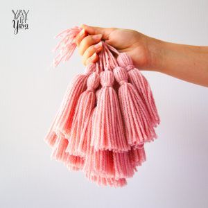 Hand holding a furls wander yarn in gray, and three skeins of furls wander yarn in brown, cream, and gray on a light wood background