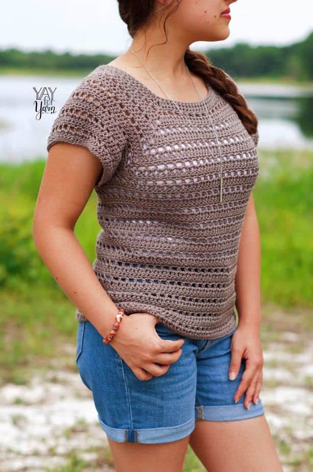 girl looking to the side, wearing brown crochet tee over white tank top with jean shorts, standing in front of lake