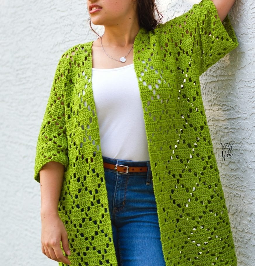 girl leaning against grey wall wearing green crocheted cardigan with white tank and jeans