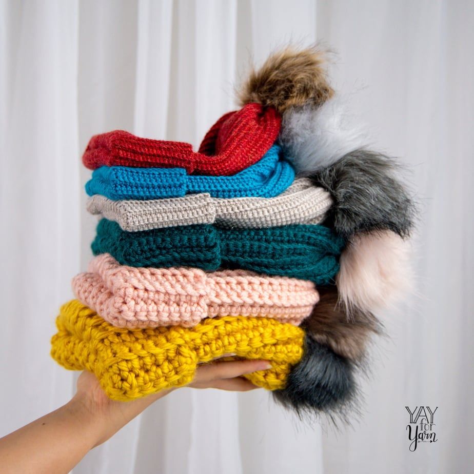 hand holding stack of crocheted hats with faux fur pom poms
