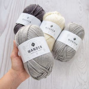 Hand holding a furls wander yarn in gray, and three skeins of furls wander yarn in brown, cream, and gray on a light wood background