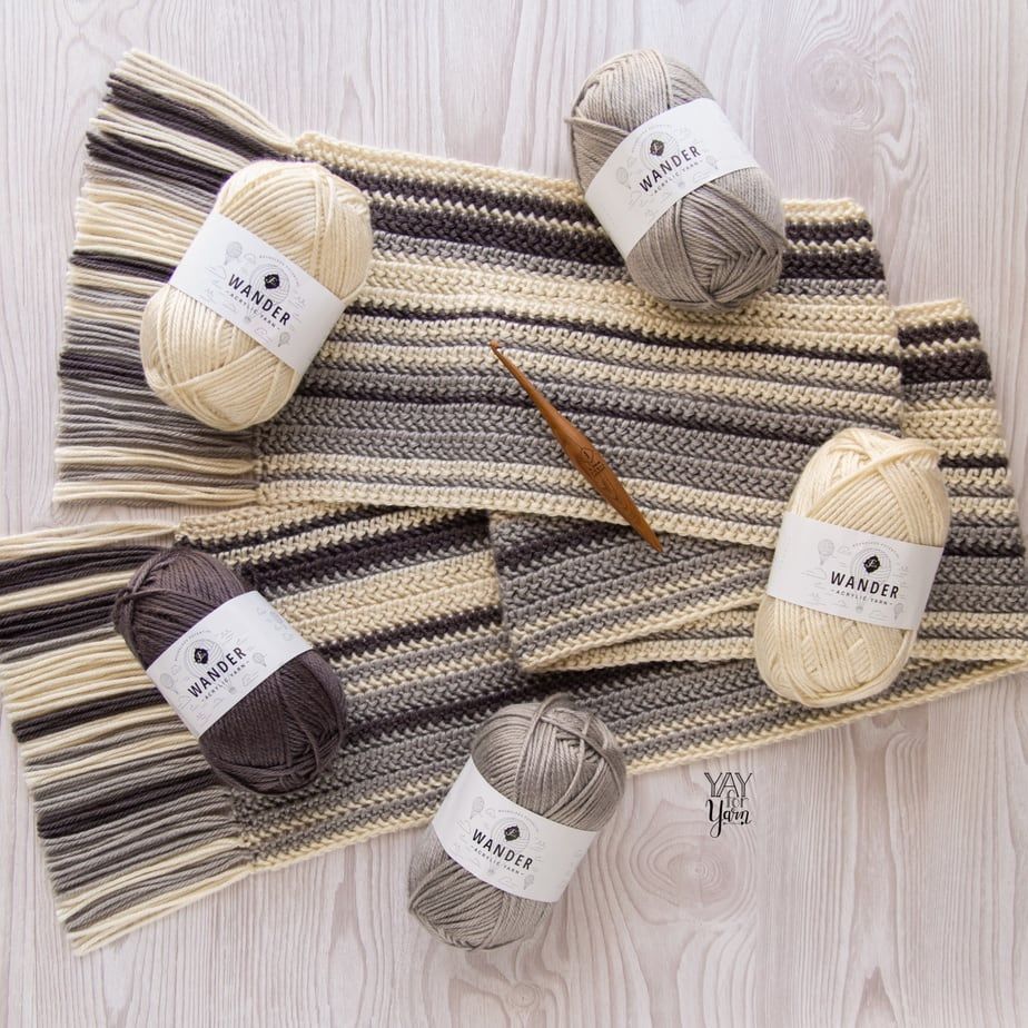 striped cream, silver, and brown scarf with fringe on wood background, with skeins of matching yarn and crochet hook