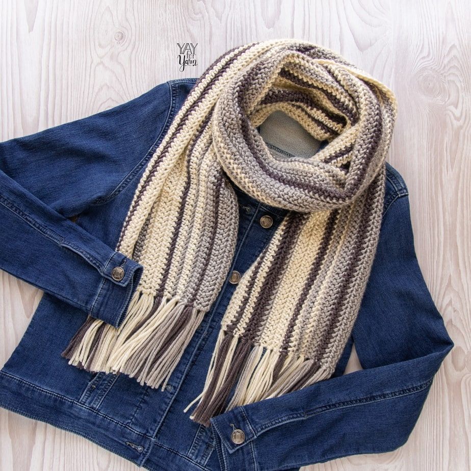 denim jacket laying on light wood with striped fringed crochet scarf in cream, grey and brown