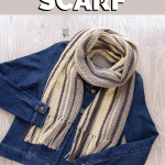denim jacket laying on light wood with striped fringed crochet scarf in cream, gray and brown with text that reads ‘Herringbone Scarf - free crochet pattern and video tutorial’