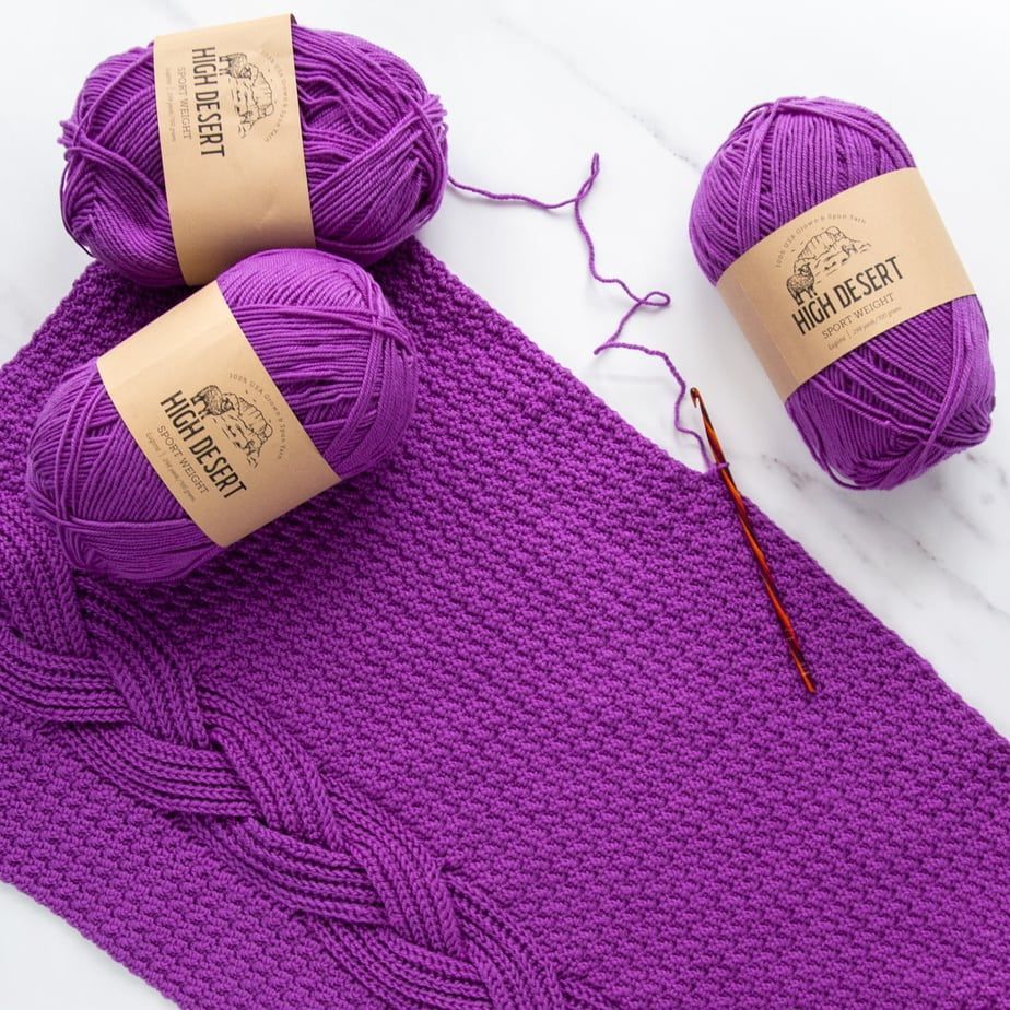 flat lay of braided cable crochet sweater in progress, purple yarn, striped hook on white marble