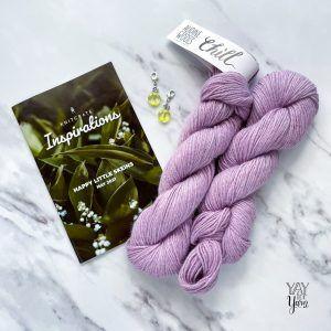 Hand holding a furls wander yarn in gray, and three skeins of furls wander yarn in brown, cream, and gray on a light wood background
